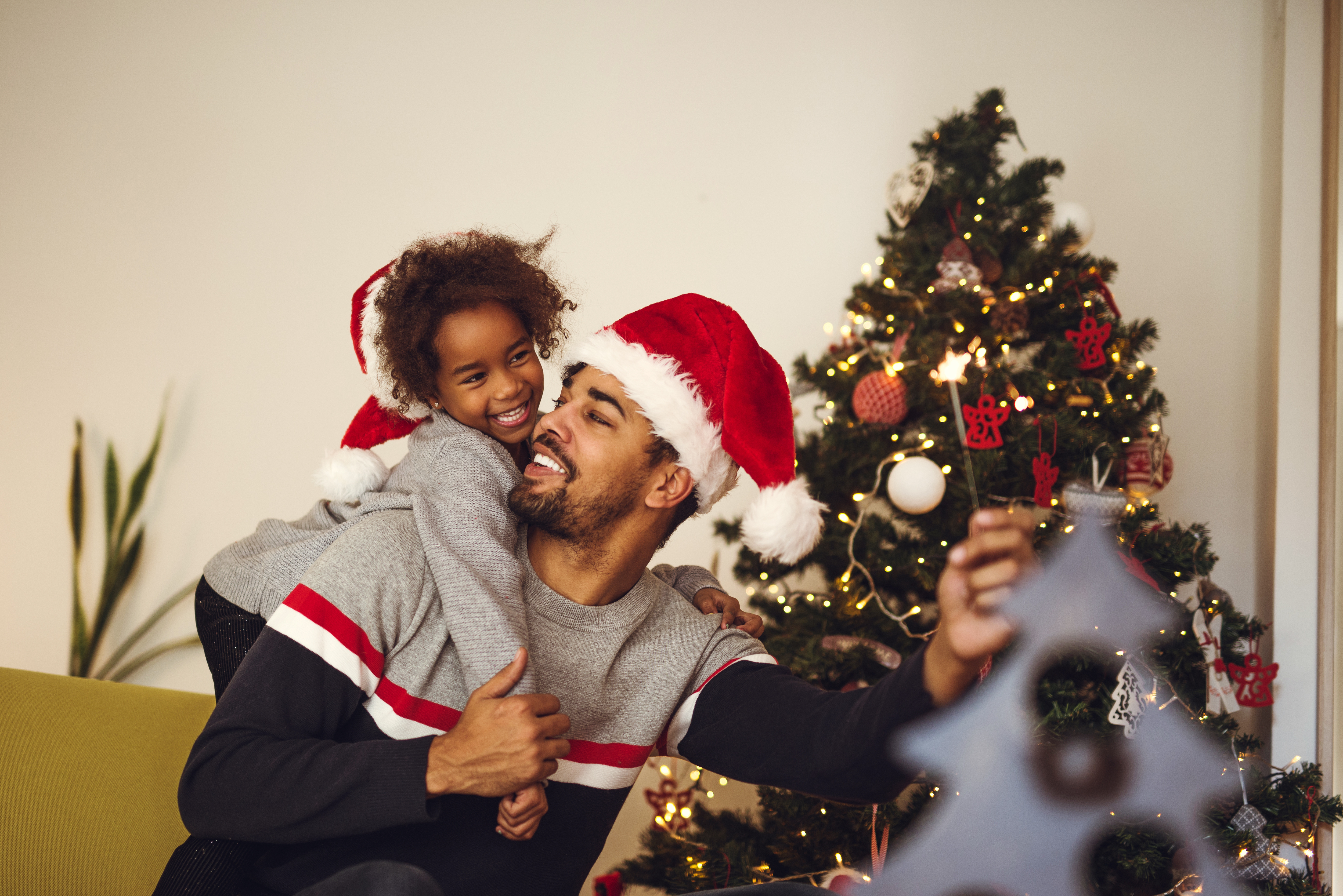 Man and child sitting together looking at one another smiling. Both are wearing Santa hats. Background features a Christmas tree decorated with ornaments.