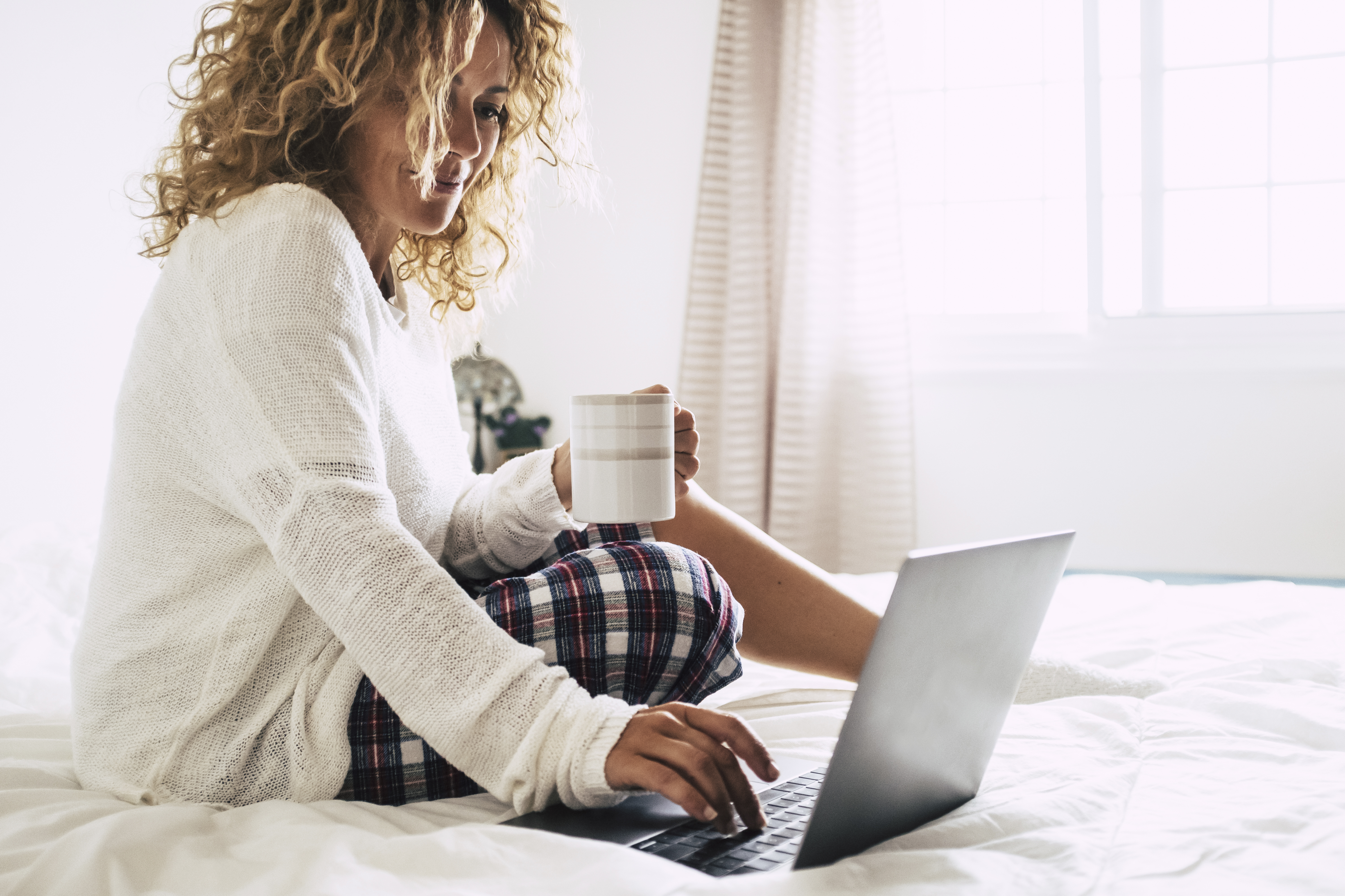 woman drinking coffee sitting on bed using laptop